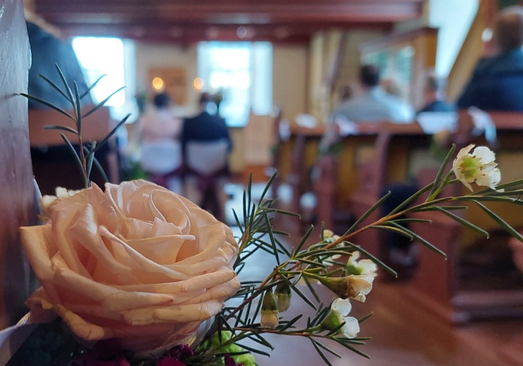 Im Vordergrund schmücken Blumen eine Hochzeit in der Kirche, in einer unscharfen Aufnahme ist eine Hochzeitszeremonie zu sehen.